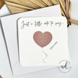 Puede incluir: Una tarjeta blanca con el texto "Just a little note to say..." y un globo rosa en forma de coraz&oacute;n con el texto "Scratch Here" debajo. La tarjeta tiene un logotipo que dice "A Maura Designs Invitation Studio".