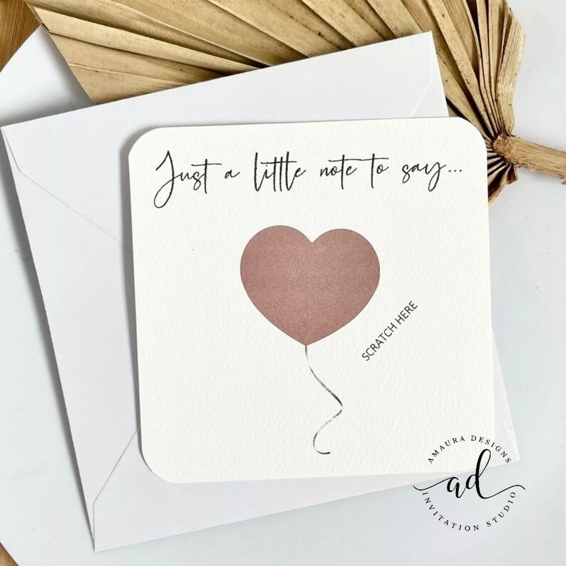 Puede incluir: Una tarjeta blanca con el texto "Just a little note to say..." y un globo rosa en forma de coraz&oacute;n con el texto "Scratch Here" debajo. La tarjeta tiene un logotipo que dice "A Maura Designs Invitation Studio".