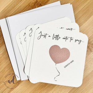 Puede incluir: Un conjunto de tarjetas blancas con un globo en forma de coraz&oacute;n rosa y el texto "Just a little note to say..." y "Scratch Here".