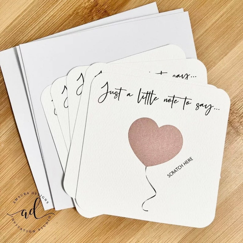 Puede incluir: Un conjunto de tarjetas blancas con un globo en forma de coraz&oacute;n rosa y el texto "Just a little note to say..." y "Scratch Here".