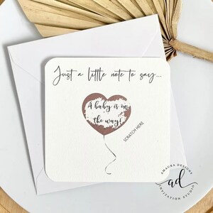 Puede incluir: Una tarjeta blanca con el texto "Just a little note to say... A baby is on the way!" con un globo en forma de coraz&oacute;n marr&oacute;n con el texto "Scratch Here" debajo del globo. La tarjeta tiene el texto "Amaura Designs Invitation Studio" en la parte inferior.