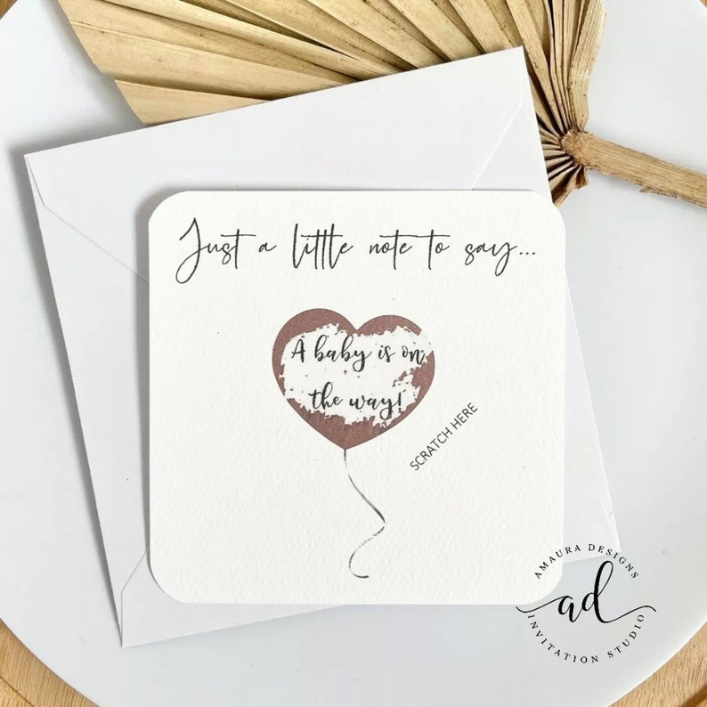 Puede incluir: Una tarjeta blanca con el texto "Just a little note to say... A baby is on the way!" con un globo en forma de coraz&oacute;n marr&oacute;n con el texto "Scratch Here" debajo del globo. La tarjeta tiene el texto "Amaura Designs Invitation Studio" en la parte inferior.