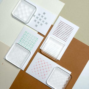 Pode incluir: Um conjunto de quatro blocos de acrílico transparente para carimbos de borracha, cada um emparelhado com um cartão branco com um padrão estampado único: uma grelha geométrica preta, um motivo floral verde, linhas castanhas e um design de ponto cruz rosa.