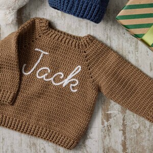 May include: A brown, crocheted sweater with the name "Jack" embroidered in white script across the front. The sweater has long sleeves and a ribbed neckline and hem. A gift box and a blue knitted hat are in the background.