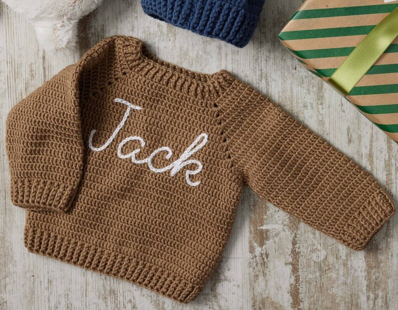 May include: A brown, crocheted sweater with the name "Jack" embroidered in white script across the front. The sweater has long sleeves and a ribbed neckline and hem. A gift box and a blue knitted hat are in the background.