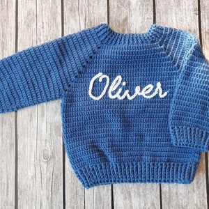 May include: A blue crochet sweater with the name "Oliver" in white script across the chest. The sweater has long sleeves and a ribbed hem and cuffs. The garment is displayed on a wooden surface.