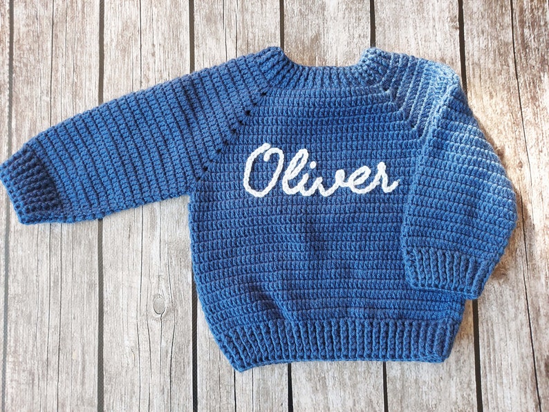 May include: A blue crochet sweater with the name "Oliver" in white script across the chest. The sweater has long sleeves and a ribbed hem and cuffs. The garment is displayed on a wooden surface.