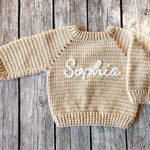 May include: A beige crochet sweater with long sleeves. The word "Sophia" is embroidered in white across the front. The sweater is made of a textured yarn and is displayed on a wooden surface.