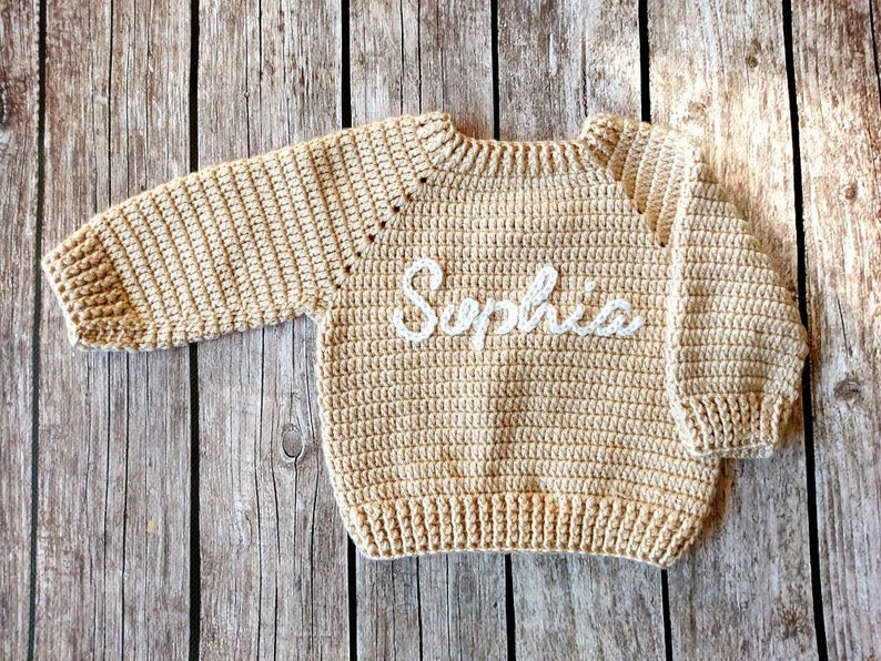 May include: A beige crochet sweater with long sleeves. The word "Sophia" is embroidered in white across the front. The sweater is made of a textured yarn and is displayed on a wooden surface.