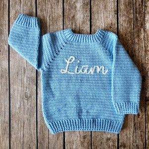 May include: A light blue, crocheted sweater with the name "Liam" embroidered in white. The sweater has long sleeves and a ribbed hem and cuffs. The background is a wooden surface.