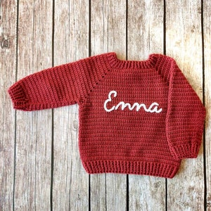 May include: A red crocheted sweater with the name "Emma" in white script across the chest. The sweater has long sleeves and a ribbed hem and cuffs. The garment is displayed on a wooden surface.