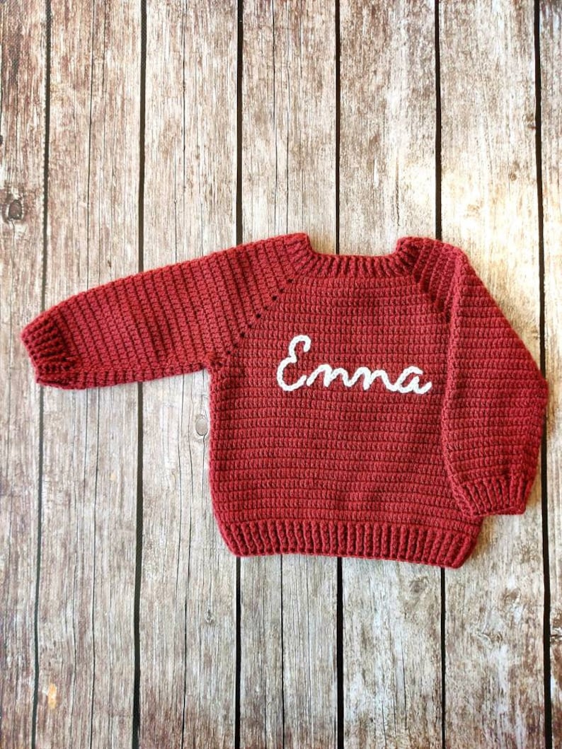 May include: A red crocheted sweater with the name "Emma" in white script across the chest. The sweater has long sleeves and a ribbed hem and cuffs. The garment is displayed on a wooden surface.