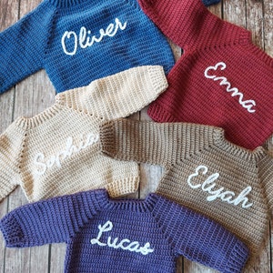 May include: A collection of handmade crochet sweaters in various colors, including blue, burgundy, beige, brown, and purple. Each sweater has a name embroidered in white script: Oliver, Emma, Sophia, Elyah, and Lucas. The sweaters are displayed on a wooden surface.