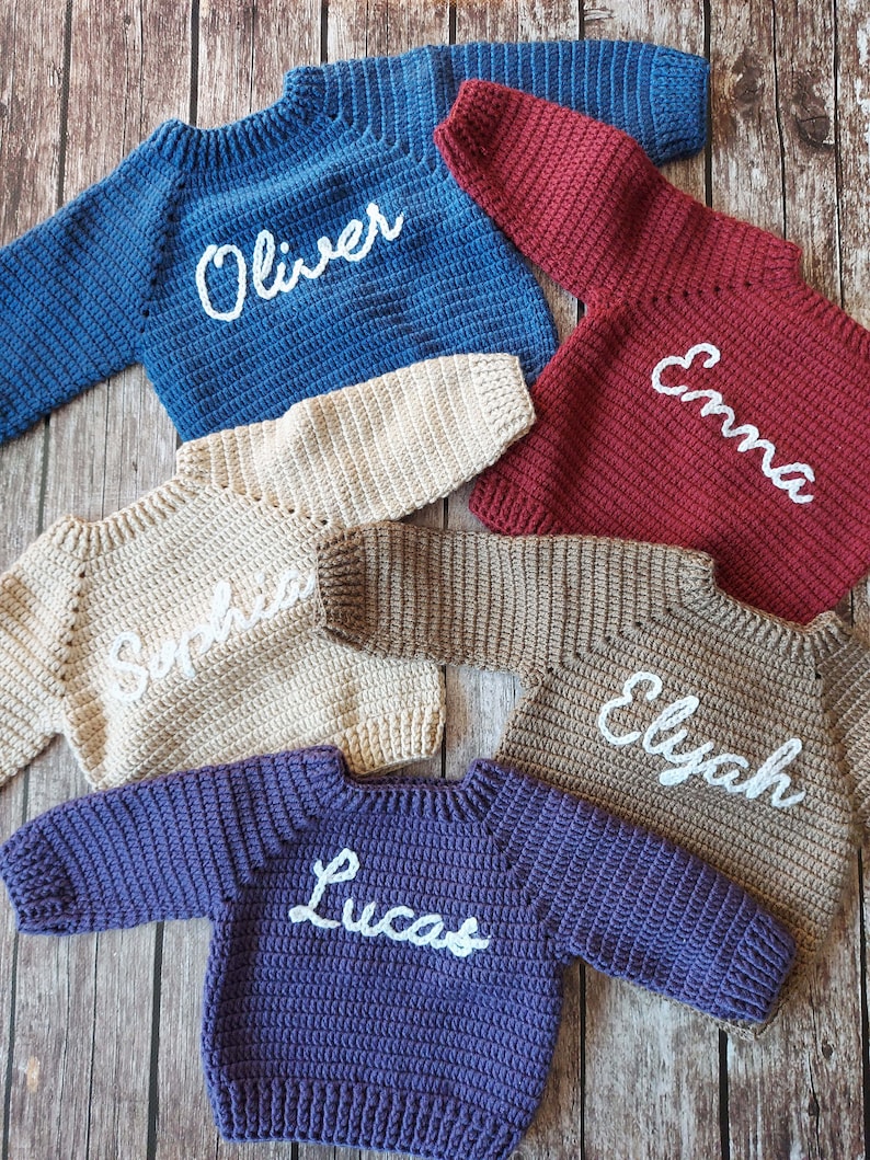 May include: A collection of handmade crochet sweaters in various colors, including blue, burgundy, beige, brown, and purple. Each sweater has a name embroidered in white script: Oliver, Emma, Sophia, Elyah, and Lucas. The sweaters are displayed on a wooden surface.