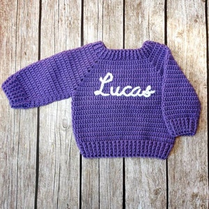 May include: A handmade, purple crochet sweater with the name "Lucas" in white script across the front. The sweater has long sleeves and a ribbed hem and cuffs. The sweater is displayed on a wooden background.