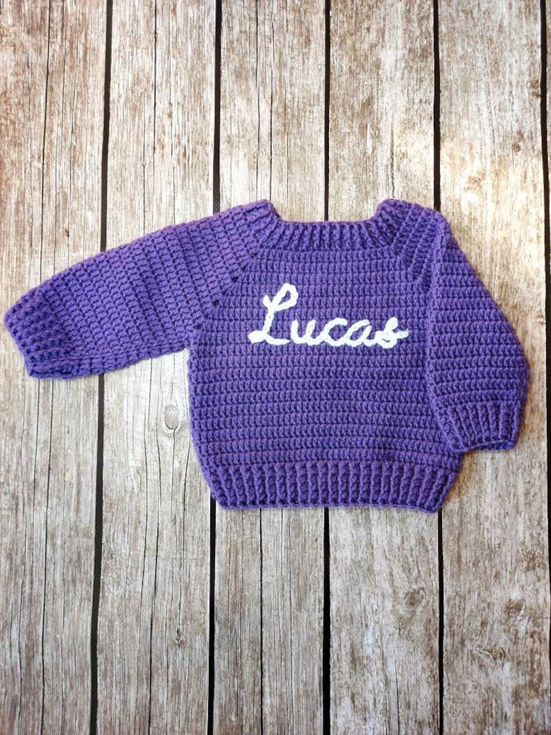 May include: A handmade, purple crochet sweater with the name "Lucas" in white script across the front. The sweater has long sleeves and a ribbed hem and cuffs. The sweater is displayed on a wooden background.