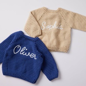 May include: Two handmade crochet sweaters. One is navy blue with the name "Oliver" embroidered in white. The other is tan with "Sophia" embroidered in white. Both sweaters have long sleeves and a crew neck.