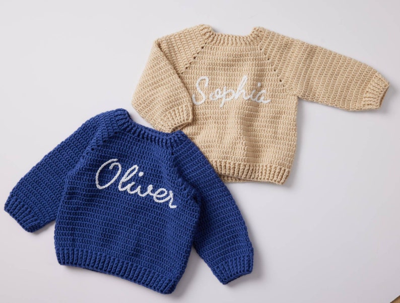 May include: Two handmade crochet sweaters. One is navy blue with the name "Oliver" embroidered in white. The other is tan with "Sophia" embroidered in white. Both sweaters have long sleeves and a crew neck.