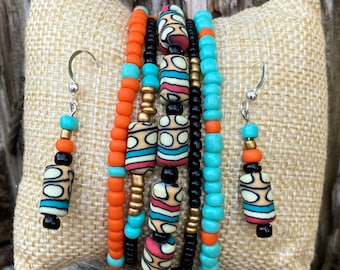 May include: A set of five colorful beaded bracelets with turquoise, orange, black, and gold beads. The bracelets are stacked on top of each other and feature larger, patterned beads. The set also includes a pair of matching earrings with turquoise, orange, and black beads.