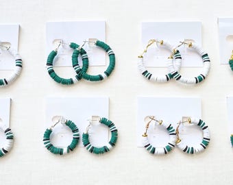 May include: Eight pairs of hoop earrings in the image. The earrings are white, green, and black, with gold or silver-colored clasps. The earrings are arranged on white cards, showcasing the jewelry's design and color variations.