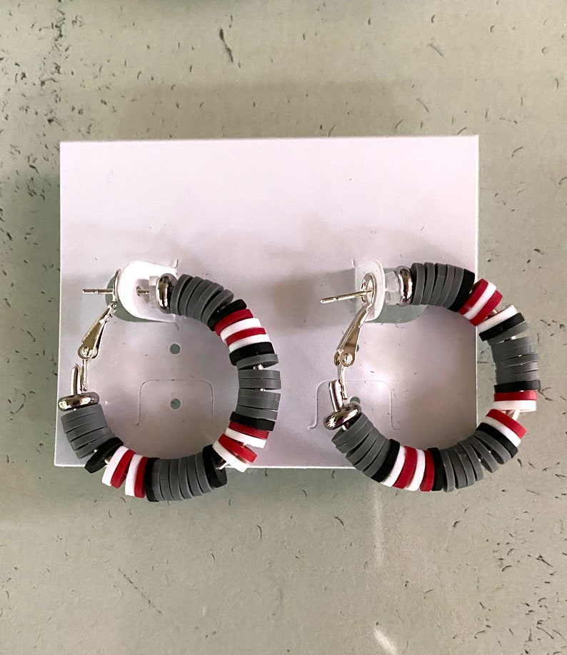 May include: A pair of hoop earrings with a gray, white, and red striped design. The earrings are made of a lightweight material and have a silver-toned finish.