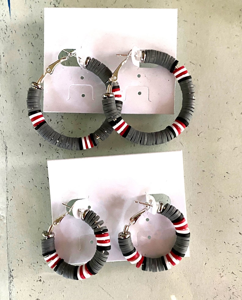 May include: Two pairs of hoop earrings with a gray, white, and red color scheme. The earrings are made of a layered material that resembles beads.