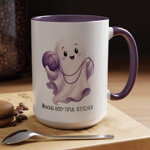 May include: A white ceramic mug with a purple interior and handle, featuring a cartoon ghost holding a purple ball of yarn. Below the ghost, the text reads "MAKING BOO-TIFUL STITCHES" in black capital letters. The mug has a smooth, cylindrical shape.