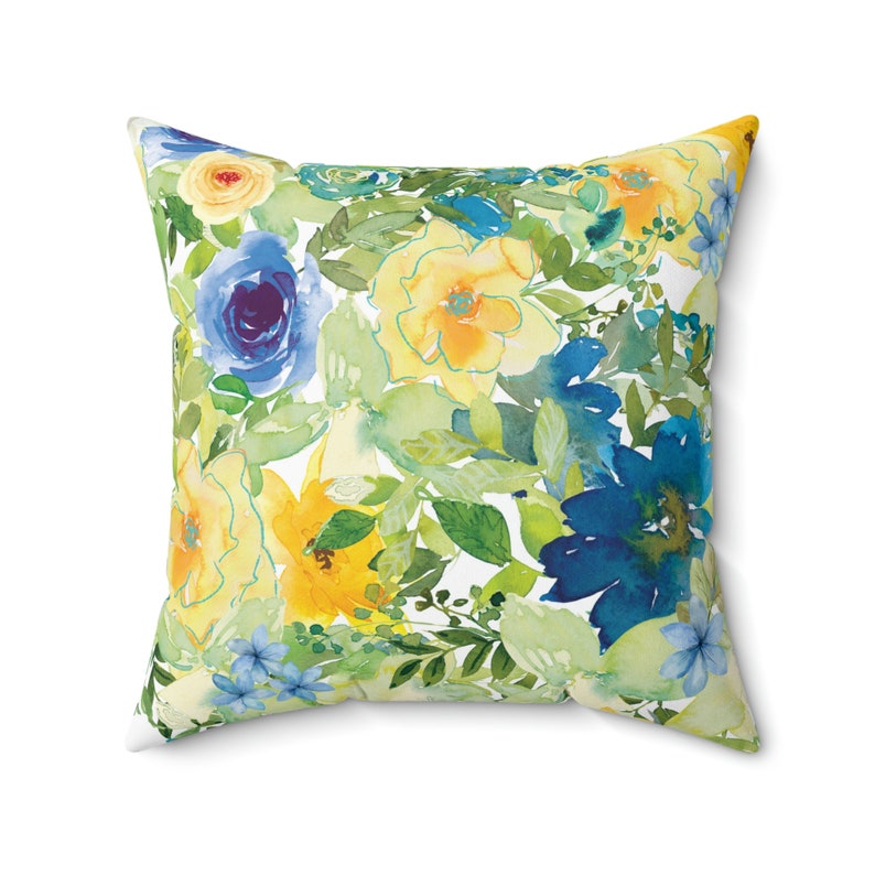 Op de afbeelding: Een vierkant kussen met een bloemenpatroon. Het kussen heeft een aquarelontwerp met gele, blauwe en groene bloemen op een witte achtergrond.