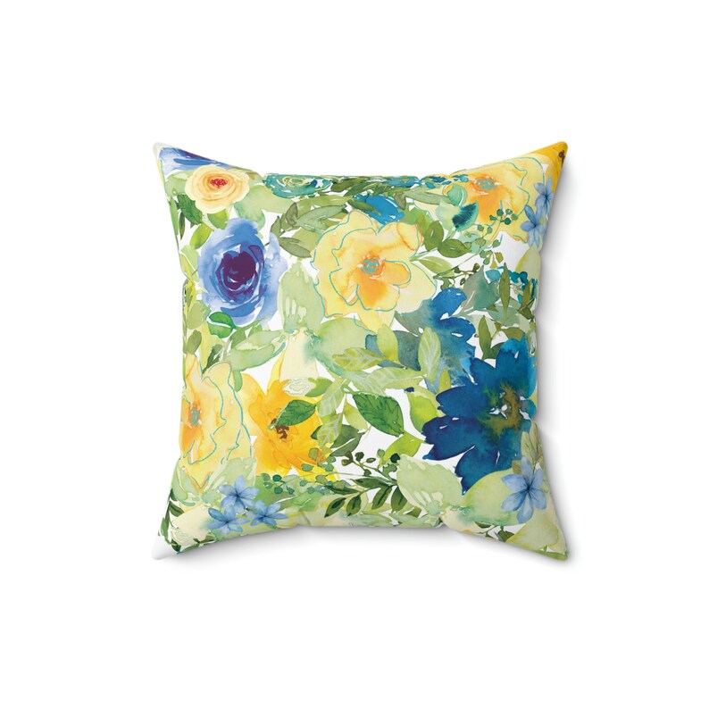 Op de afbeelding: Een vierkant kussen met een bloemenpatroon. Het kussen heeft een aquarelontwerp met gele, blauwe en groene bloemen op een witte achtergrond.