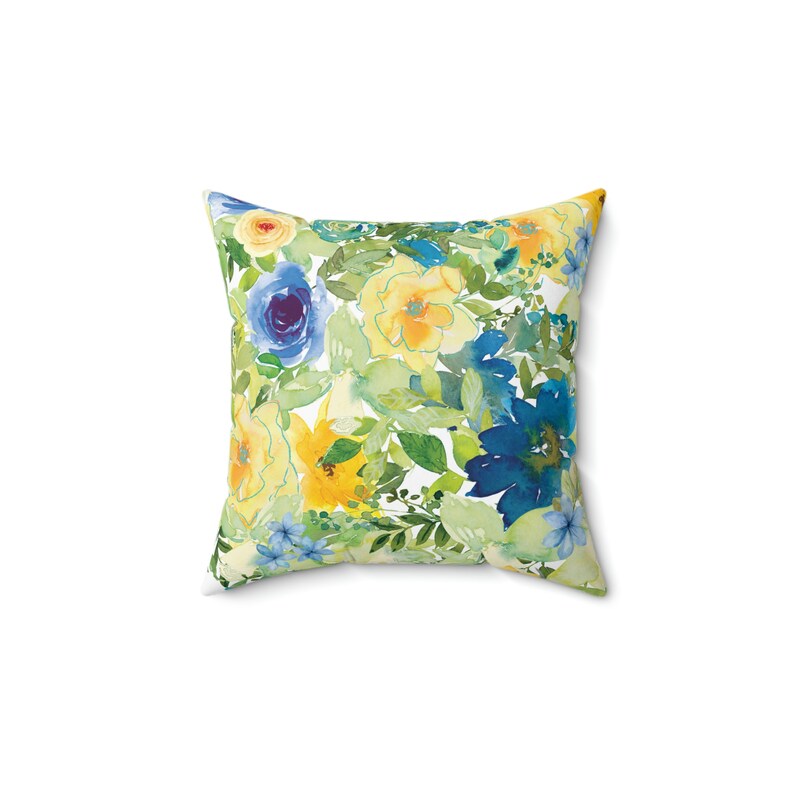 Op de afbeelding: Een vierkant kussen met een bloemenpatroon. Het kussen heeft een aquarelontwerp met blauwe, gele en groene bloemen op een witte achtergrond.