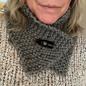 May include: A gray knitted cowl neck scarf with a black plastic clasp. The scarf is made of a chunky yarn and has a textured look.