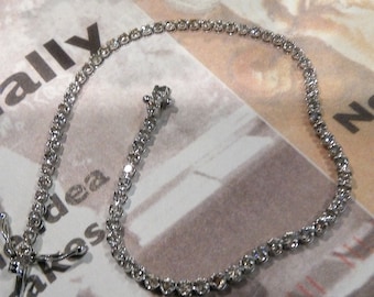 Puede incluir: Un brazalete de tenis plateado con pequeñas piedras transparentes. El brazalete está sobre una superficie blanca con un fondo borroso.