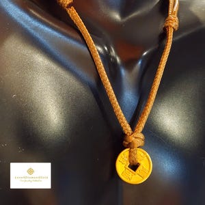 Puede incluir: Un collar con colgante dorado con un cordón marrón. El colgante circular tiene un agujero cuadrado y caracteres chinos. El cordón está anudado en la parte superior e inferior del colgante. El collar se exhibe en un maniquí negro.