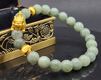 May include: A jade bead bracelet with gold accents and a gold figurine. The bracelet features light green jade beads and gold-colored beads. A gold figurine of a mythical creature is also present. The bracelet is displayed on a dark surface.