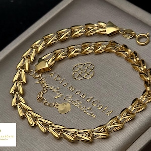 Op de afbeelding: Een gouden kettingarmband met een herhalend bladachtig schakelpatroon. De armband is voorzien van een ronde veerringsluiting en een verstelbare verlengketting met een kleine hartvormige bedel van gepolijst metaal.