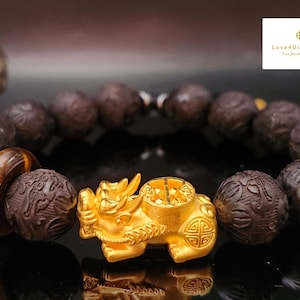 May include: A beaded bracelet featuring dark brown carved beads, a tiger's eye bead, and a gold-colored Pixiu charm. The Pixiu is a mythical creature, and the bracelet is likely a symbol of wealth and good fortune.