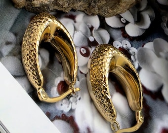 May include: A pair of gold-colored hoop earrings with a textured, diamond-like pattern. The earrings have a curved, oval shape and a hinged closure. The background features a floral pattern and a rock.
