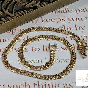 Puede incluir: Un collar de cadena de oro con un diseño de eslabones planos entrelazados en forma de espiga. El collar incluye un mecanismo de cierre de caja rectangular con un pestillo de seguridad. El metal tiene un acabado pulido y reflectante.