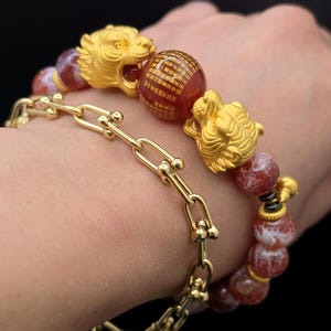 May include: A gold chain link bracelet paired with a beaded bracelet featuring two gold tiger head charms and a central red bead inscribed with gold Chinese characters. The beaded bracelet consists of round, marbled red and white stone beads.