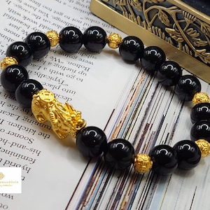 May include: A beaded bracelet featuring large, glossy black beads interspersed with smaller gold-colored beads. A detailed gold-colored charm is also present. The bracelet is displayed on a printed surface.