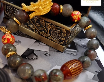 May include: A beaded bracelet featuring a gold dragon head and various gemstone beads in shades of gray, red, and gold. The bracelet includes a red bead with gold accents and is displayed on a decorative box. The text "Love4DiamondGold" is visible.