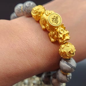 以下が含まれることがあります： 「福」の漢字が刻まれた、Pixiuのチャームが付いたゴールドのブレスレット。グレーと白の大理石模様の石ビーズとゴールドのアクセントビーズが含まれています。手首に着用されています。