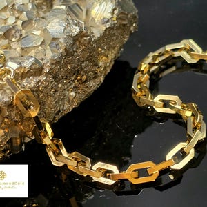 May include: A gold chain bracelet with a geometric link design. The bracelet is displayed on a dark surface with a textured, gold-colored rock in the background. The bracelet is made of gold-colored metal and has a lobster clasp.
