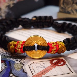 Puede incluir: Un brazalete trenzado negro con un dije de moneda china de oro y detalles rojos y dorados. La moneda tiene caracteres chinos.