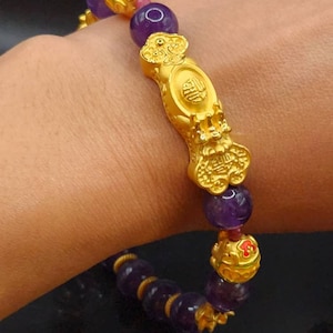 May include: A gold bracelet with a dragon charm and amethyst beads. The bracelet is on a person's wrist.