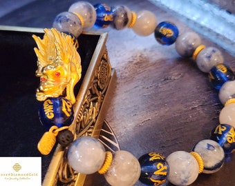 May include: A beaded bracelet featuring a gold dragon charm with red eyes and blue and white beads. The bracelet has gold accents and is displayed on a decorative gold surface. The beads have gold Chinese characters.