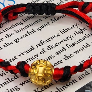 Pode incluir: Um bracelete de corda trançada vermelho e preto com um amuleto dourado com um design circular. O bracelete está sobre uma superfície branca com texto.