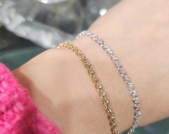Puede incluir: Dos delicados brazaletes de diamantes, uno en oro amarillo y otro en oro blanco, en la muñeca de una persona. Ambos brazaletes están hechos de una cadena fina con pequeños diamantes redondos engastados en cada eslabón. Los brazaletes son una opción popular para las mujeres que buscan un look simple y elegante.
