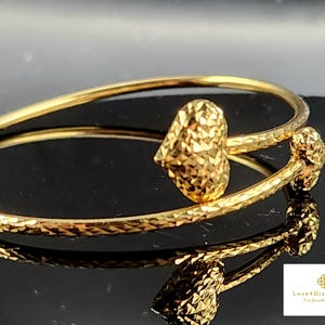 Könnte beinhalten: Ein goldfarbenes Armband mit strukturierter Oberfläche. Das Armband hat zwei dekorative Elemente mit facettiertem Design. Es wird auf einer reflektierenden Oberfläche präsentiert, die die Details hervorhebt. Ein elegantes Schmuckstück.