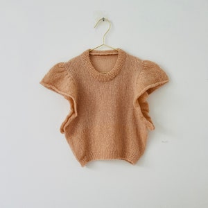 May include: A light brown knitted sleeveless top with ruffled sleeves hanging on a gold hanger against a white wall.